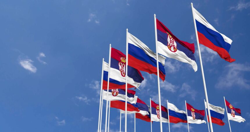 Multiple Serbian and Russian flags waving against a blue sky, symbolizing diplomatic relations and political ties between Serbia and Russia.